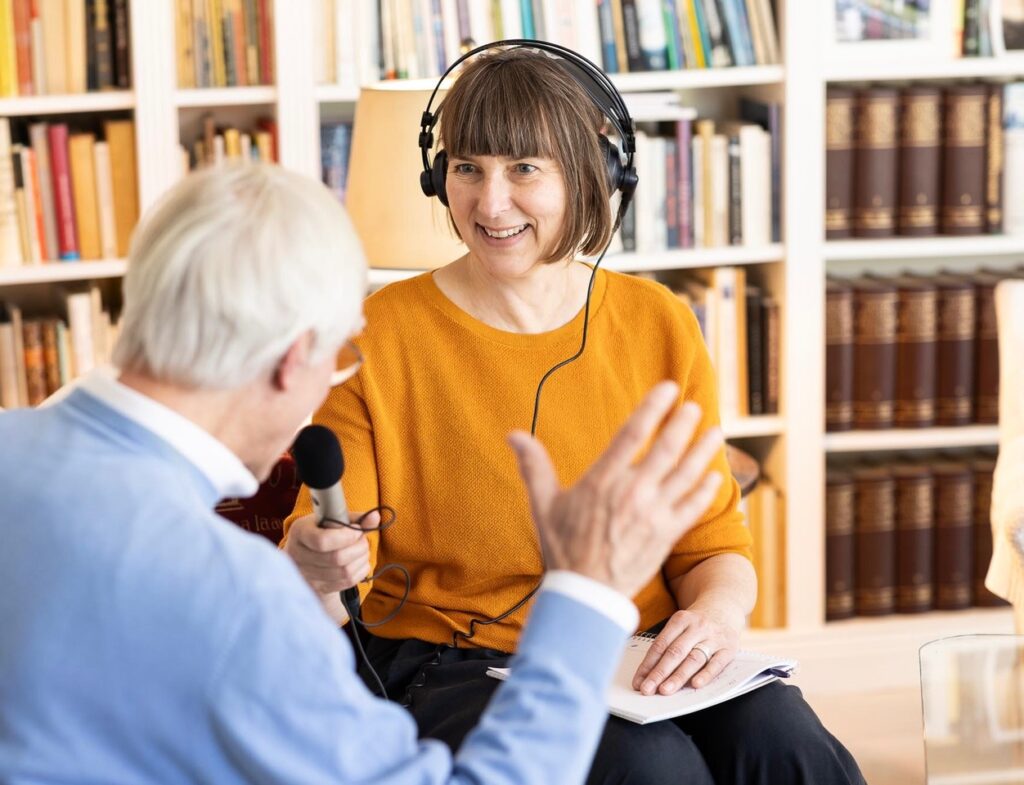 Ich sitze vor einer Bücherwand, habe professionelle Kopfhörer auf und ein Mikrofon in der Hand. Mir gegenüber sitzt mit dem Rücken zu den Betrachtenden ein gestikulierender älterer Mann, der mir offenbar etwas erzählt. Ich höre aufmerksam lächelnd zu.